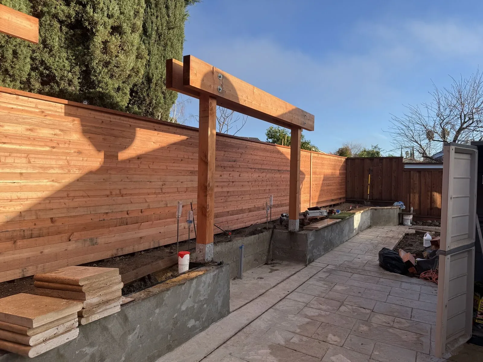Timber posts and beams being installed above new concrete retaining walls and a stamped patio in the Hernandez backyard.