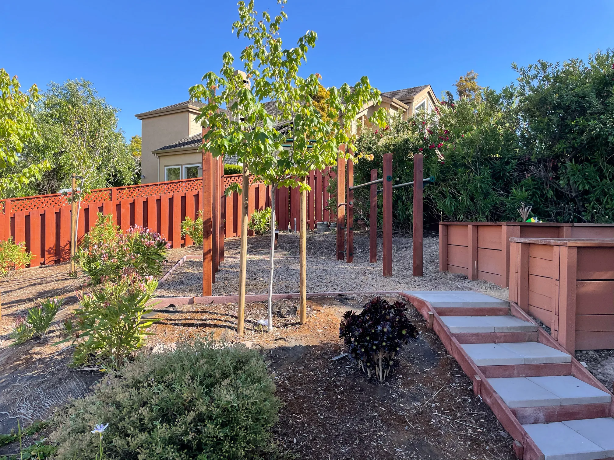 A landscaped upper garden with young trees, exercise bars, raised planters, and steps leading between levels.